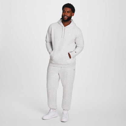 A man stands against a white background, wearing a Champion Powerblend Hoodie, C Logo - Big & Tall in light gray with matching sweatpants and white sneakers. His left hand is in his pocket and he smiles slightly.