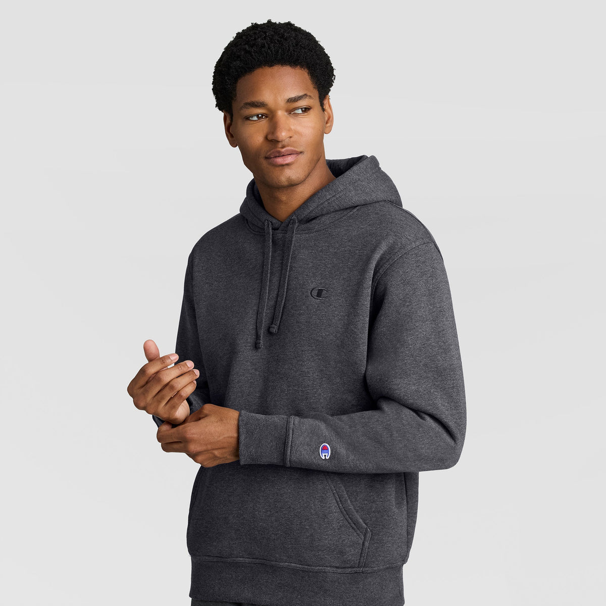 A man wearing a Champion Powerblend Hoodie, C Logo in dark gray stands against a plain light gray background, looking slightly to his left with one hand raised near his chest.