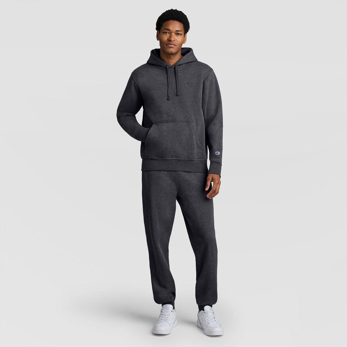 A man stands facing forward, wearing the Champion Powerblend Hoodie, C Logo in dark gray with matching sweatpants and white sneakers, against a plain light gray background.