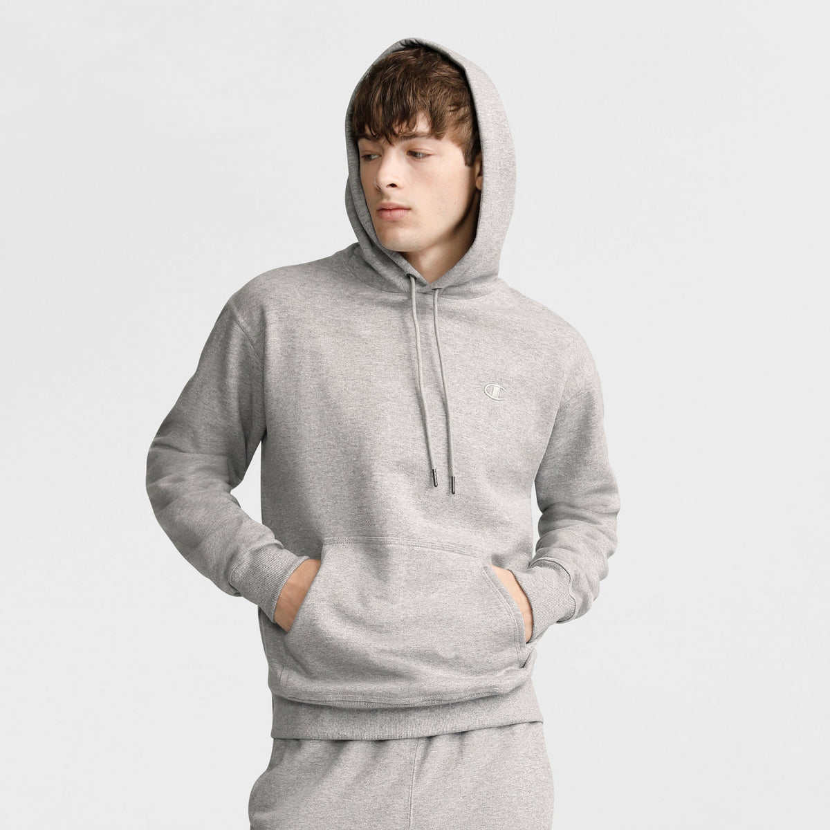 Young person in a light gray Champion Powerblend Hoodie, C Logo and matching sweatpants stands with hands in front pocket, looking to the side against a plain light background.