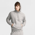 Young person in a light gray Champion Powerblend Hoodie, C Logo and matching sweatpants stands with hands in front pocket, looking to the side against a plain light background.