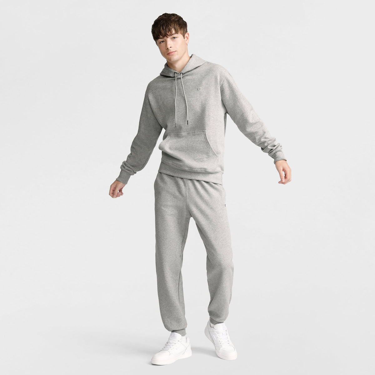 Wearing the Champion Powerblend Hoodie, C Logo in light gray with matching sweatpants and white sneakers, a person stands against a plain white background, looking at the camera.