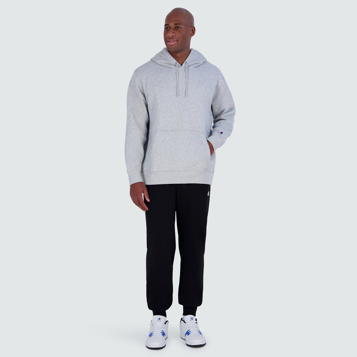 A man wearing a light gray Champion Powerblend Hoodie, C Logo - Big & Tall with a front pocket, black sweatpants, and white sneakers.