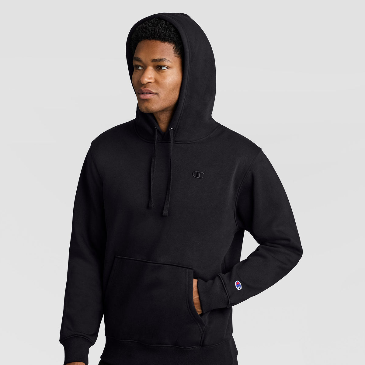 A person wearing a black Champion Powerblend Hoodie, C Logo, with the hood up stands against a plain light background, one hand casually tucked in the front pocket and a neutral expression.
