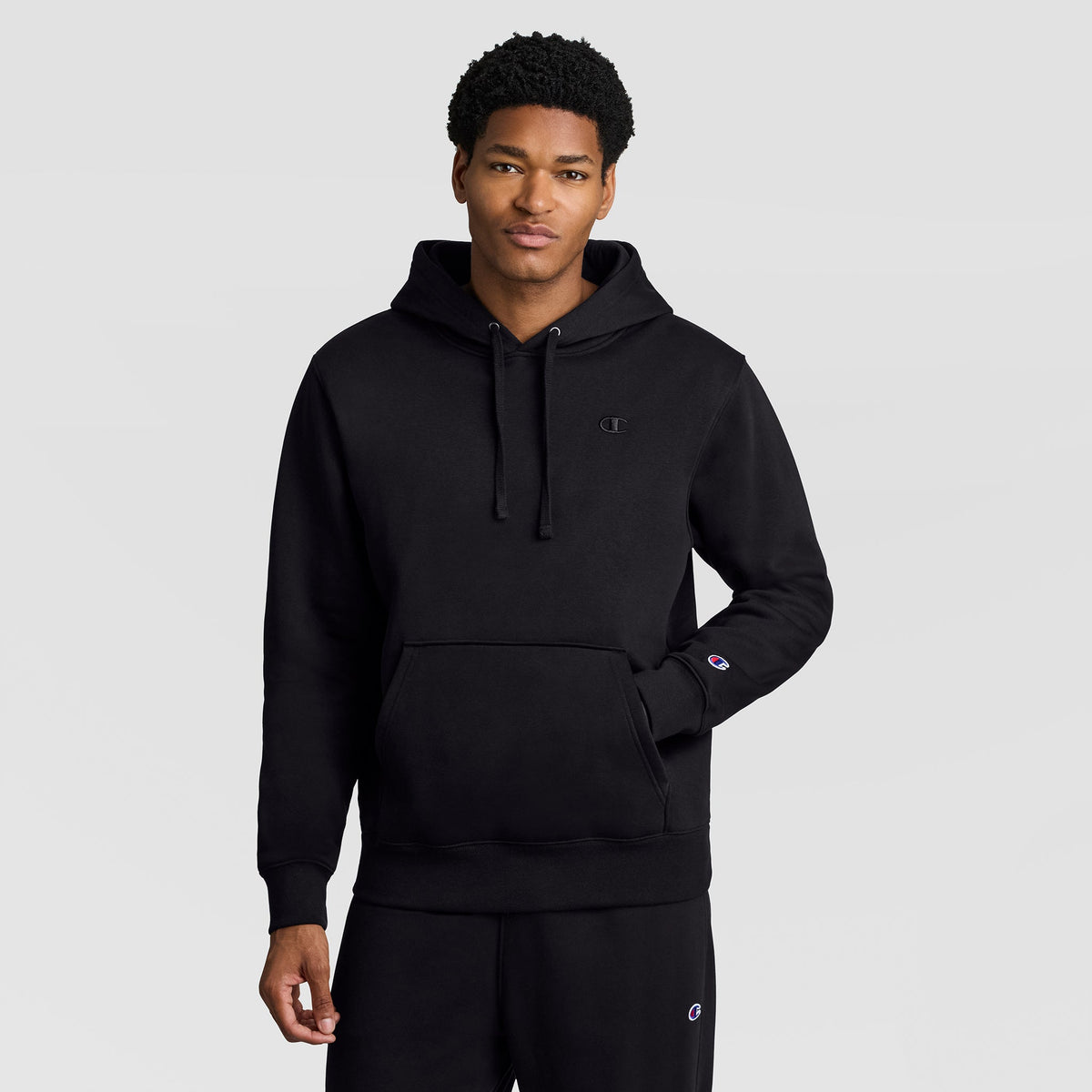 A person wearing a Champion Powerblend Hoodie, C Logo in black and black sweatpants stands against a plain light gray background, with one hand in the front pocket of the men’s hooded sweatshirt.