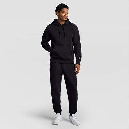 A man stands slightly turned to the side against a plain light gray background, wearing a matching black Champion Powerblend Hoodie, C Logo and sweatpants set with white sneakers.