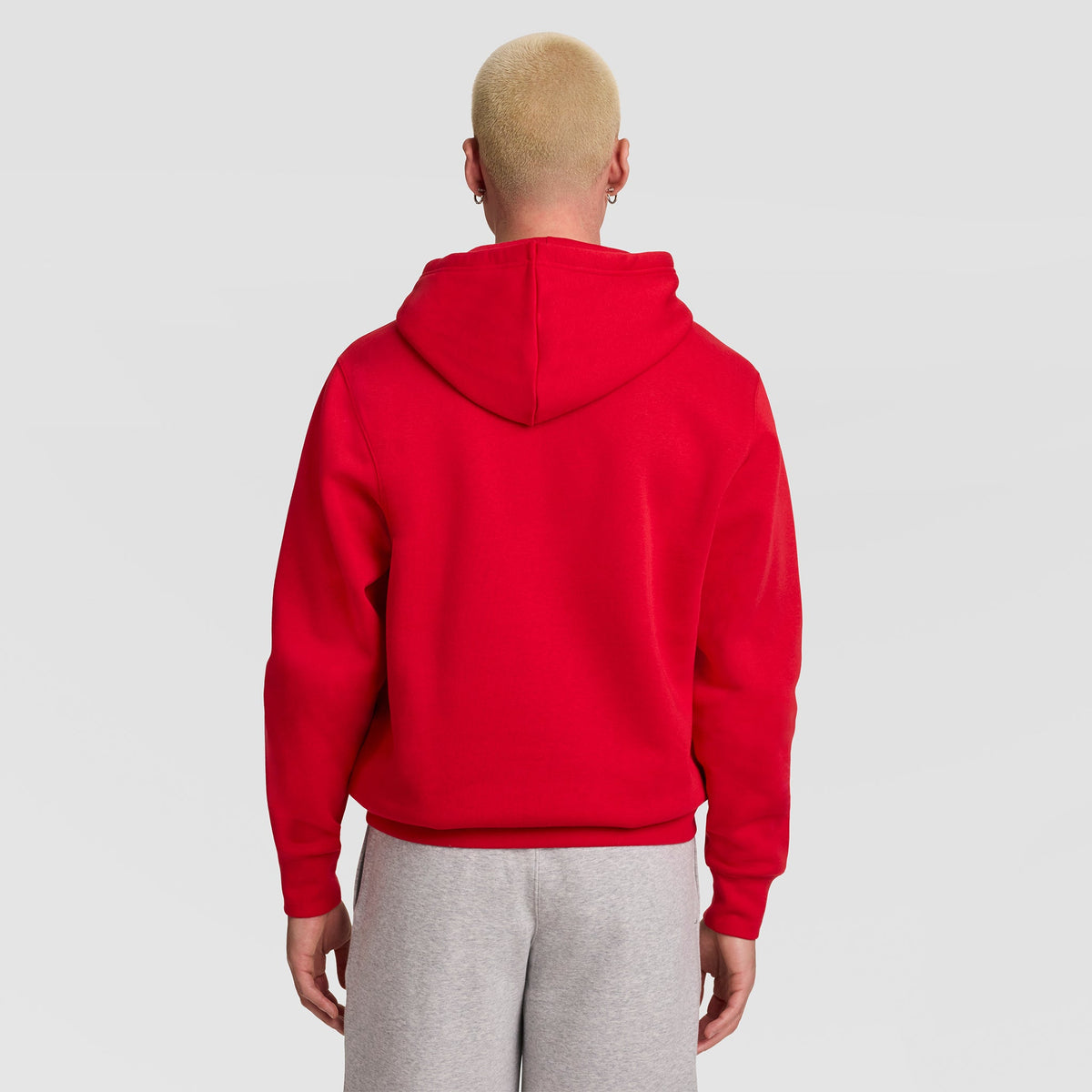 A person with short blond hair is shown from behind, wearing a bright red Champion Powerblend Hoodie, C Logo, and light gray sweatpants, standing against a plain light gray background.