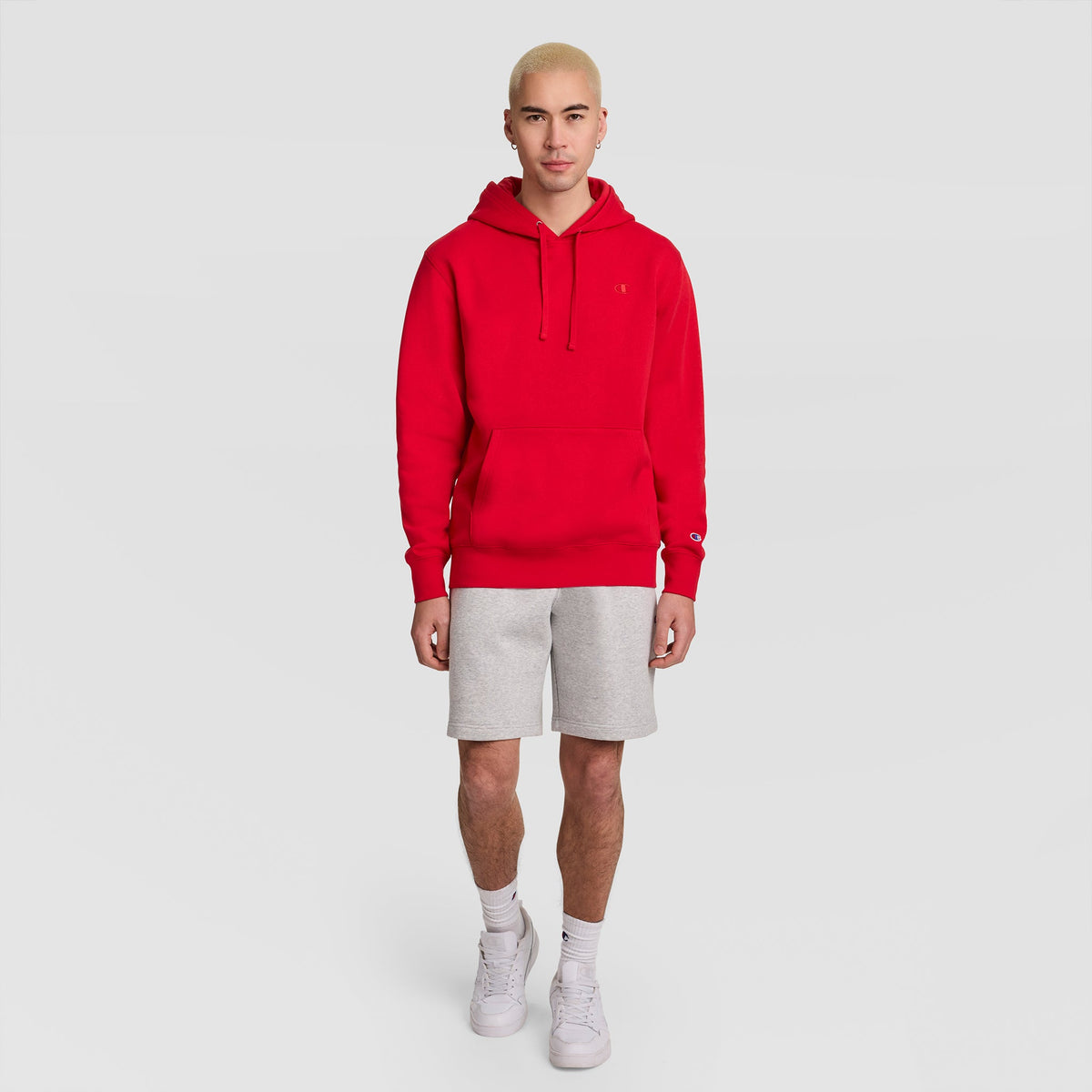 A person wearing a red Champion Powerblend Hoodie, C Logo and gray shorts stands against a plain background.