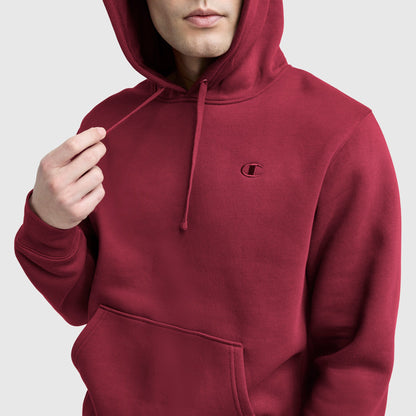 A person in a red Champion Powerblend Hoodie, C Logo with front pocket and drawstrings adjusts the hood. The C logo is visible on the left chest against a plain, light-colored background.
