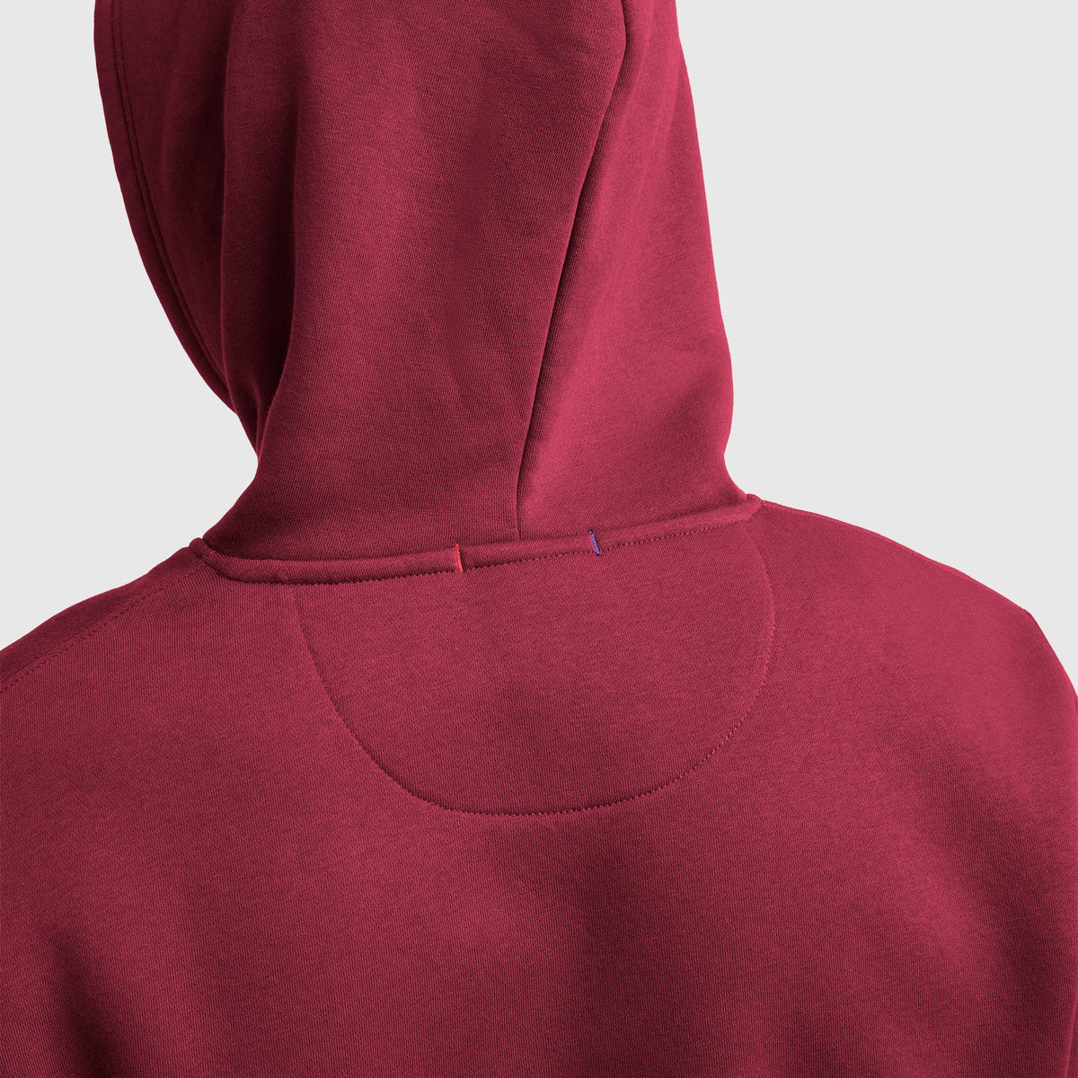 A close-up of the back of a maroon Powerblend Hoodie, C Logo by Champion shows detailed stitching and two small colored stitches—one red, one blue—on the seam below the hood.