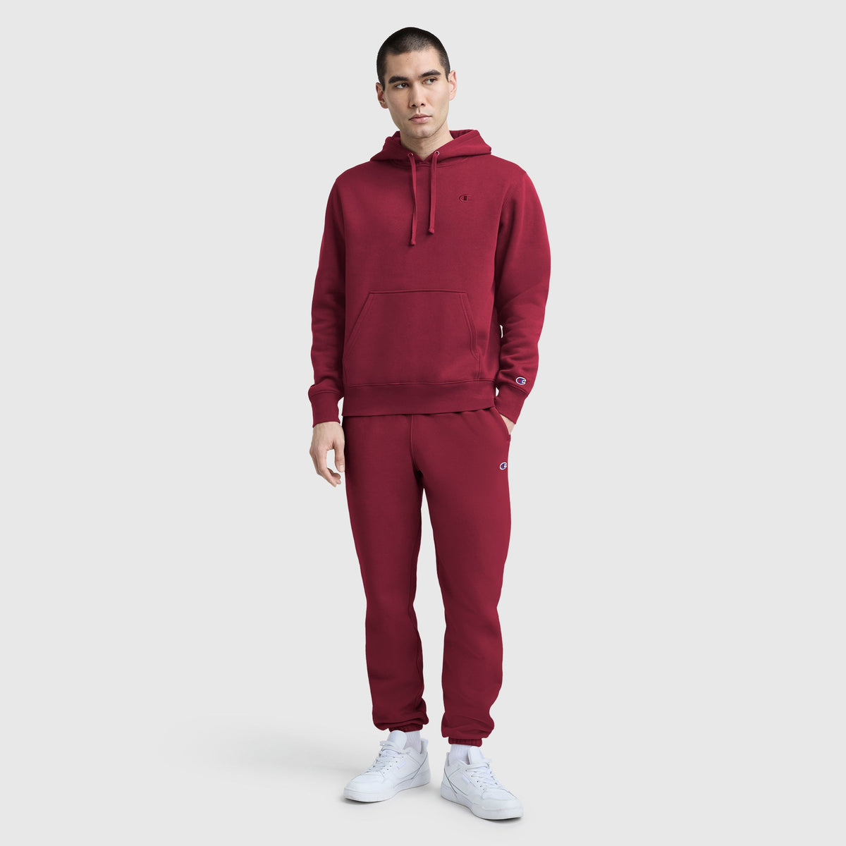 A man in a matching maroon Champion Powerblend Hoodie, C Logo and sweatpants with white sneakers stands against a plain light gray background.