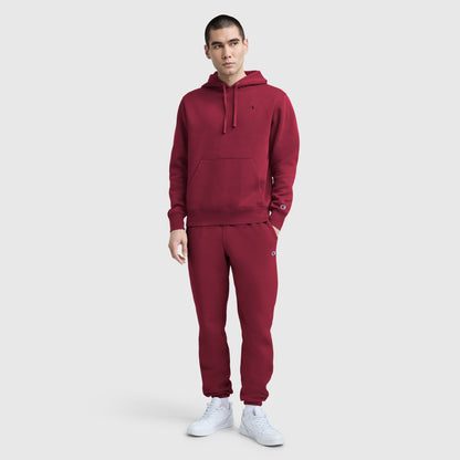 A man in a matching maroon Champion Powerblend Hoodie, C Logo and sweatpants with white sneakers stands against a plain light gray background.