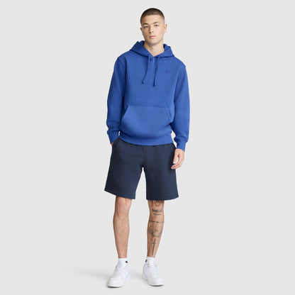 A person wearing the Champion Powerblend Hoodie, C Logo in blue, paired with dark shorts and white sneakers, stands against a plain background. Their hands rest in their pockets and one leg is slightly bent, highlighting this men’s classic hooded sweatshirt from Champion with effortless style.