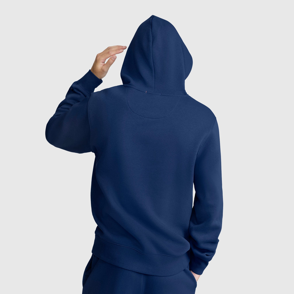 A person in a navy blue Powerblend Hoodie, C Logo by Champion and matching pants stands facing away, adjusting the hood against a plain light gray background.