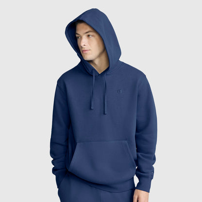 A person wears a blue Champion Powerblend Hoodie, C Logo with the hood up, front pocket, and drawstrings, standing against a plain light gray background.