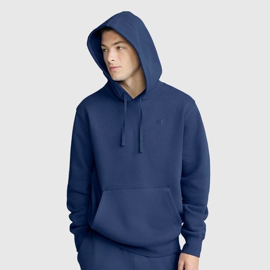 A person wears a blue Champion Powerblend Hoodie, C Logo with the hood up, front pocket, and drawstrings, standing against a plain light gray background.