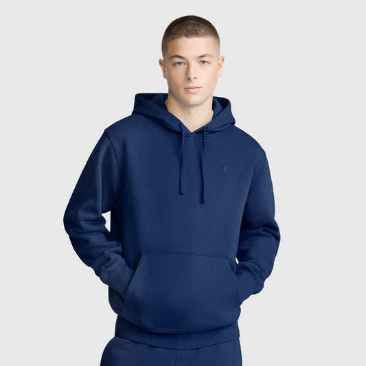 A person wearing a plain navy blue Champion Powerblend Hoodie, C Logo and matching sweatpants stands with hands in the hoodie pocket against a light gray background.