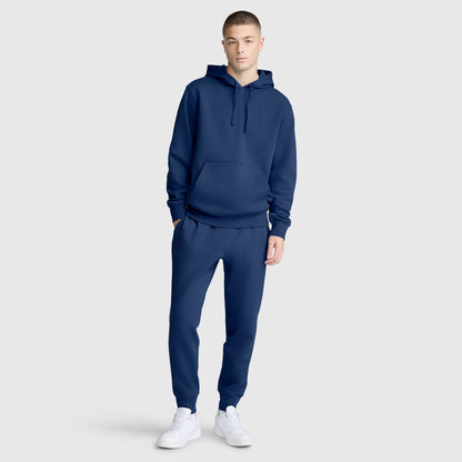 A young person in a matching navy blue Champion Powerblend Hoodie, C Logo and jogger pants stands against a plain light gray background, hands in pockets and wearing white sneakers.