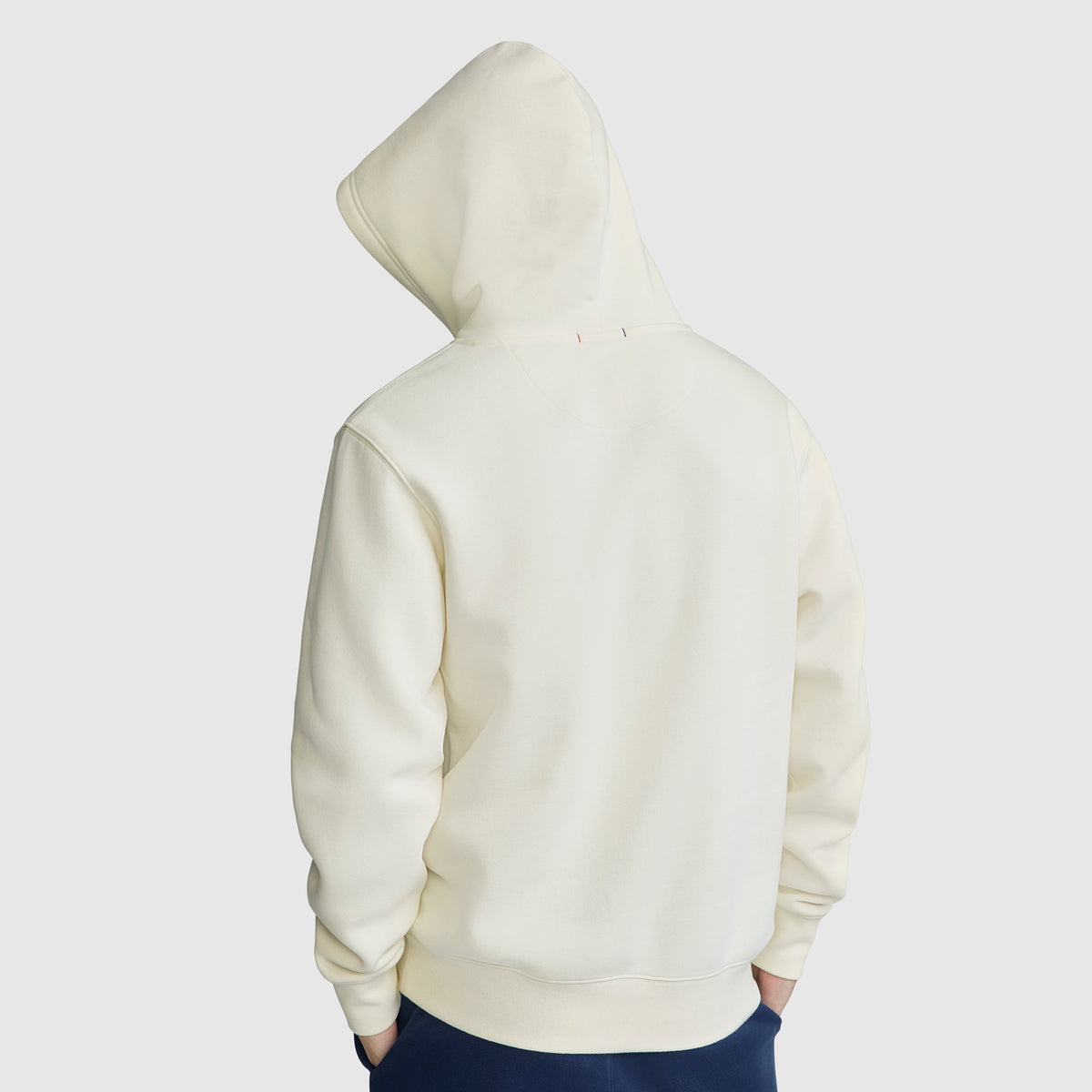 A person is wearing a Champion Powerblend Hoodie, C Logo, in off-white with the hood up, facing away from the camera. The men’s hooded sweatshirt appears plain, and the individual has their hands in their pockets. The background is light gray.