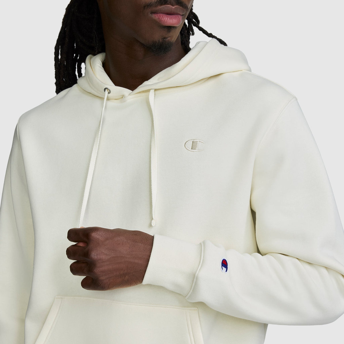 A person wearing a cream-colored Champion Powerblend Hoodie, C Logo—an ultra-comfortable fleece hoodie for men with the signature small Champion logo on the chest and sleeve—holding the hoodie with one hand.