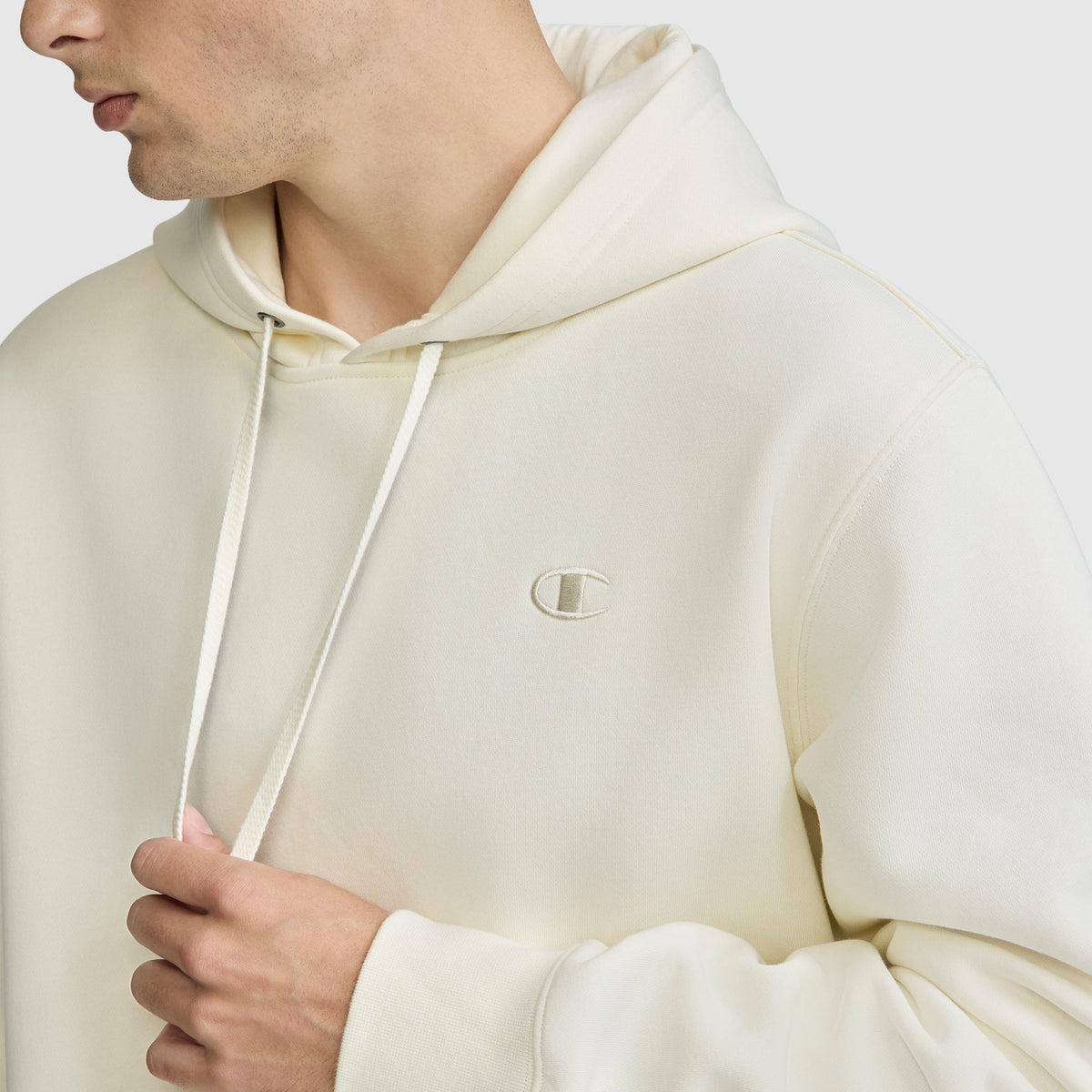 A person wearing a Champion Powerblend Hoodie, C Logo in off-white holds the drawstring with one hand. The upper body and part of the face are visible, showcasing the embroidered Champion logo on the chest. The background is plain light gray.