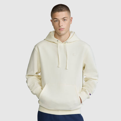 A person wearing a cream-colored Champion Powerblend Hoodie, C Logo, featuring a front pocket and drawstrings, stands facing forward with hands in the pocket against a plain light gray background.
