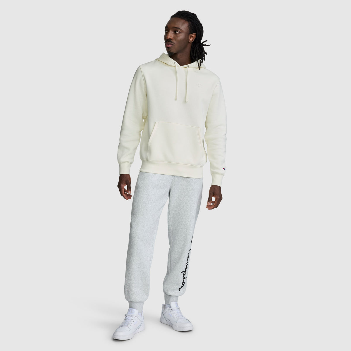 A person wearing a cream-colored Champion Powerblend Hoodie, C Logo for men, light gray sweatpants with text on one leg, and white sneakers stands against a plain light gray background.