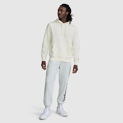 A person wearing a cream-colored Champion Powerblend Hoodie, C Logo for men, light gray sweatpants with text on one leg, and white sneakers stands against a plain light gray background.