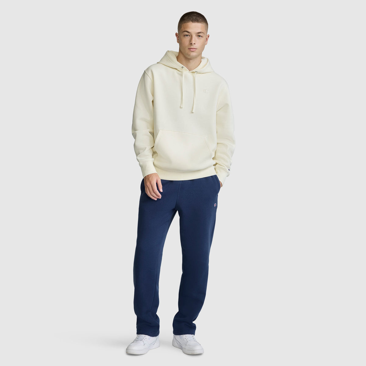 A person stands against a plain background, wearing a cream Champion Powerblend Hoodie with the C Logo, navy blue sweatpants, and white sneakers, looking at the camera with hands in pockets.