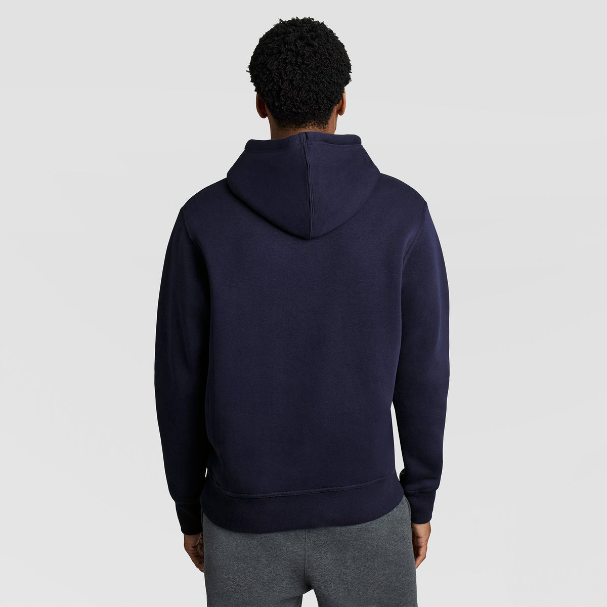 A person stands facing away against a light gray background, wearing gray pants and a dark blue Champion Powerblend Hoodie with the C Logo.