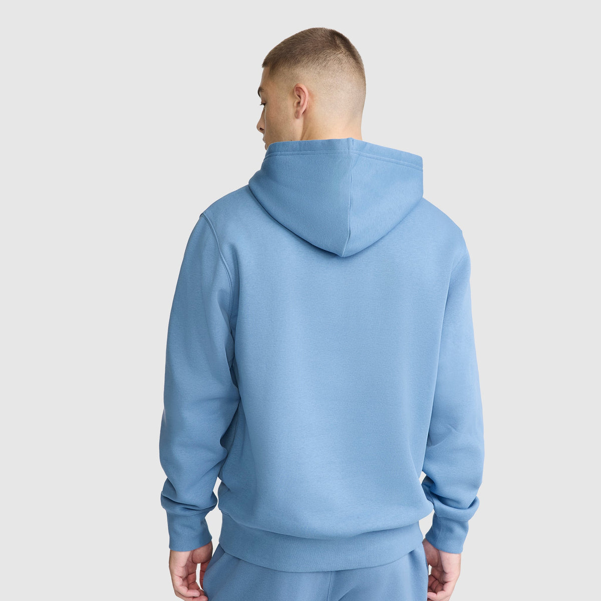 A person wearing a light blue Champion Powerblend Hoodie, C Logo, along with matching sweatpants, is standing with their back to the camera against a plain white background.