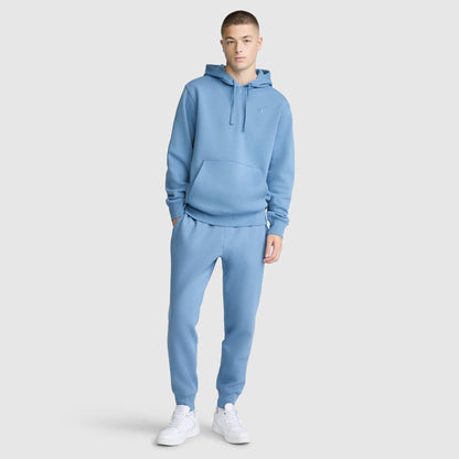 A person stands against a plain light gray background wearing a matching Champion Powerblend Hoodie, C Logo, and jogger pants in light blue, paired with white sneakers.