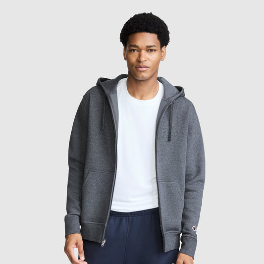 A man wearing a Champion Powerblend Full-Zip Hoodie, C Logo in dark gray over a white T-shirt and navy Powerblend cinched bottom fleece sweatpants stands against a plain light gray background.