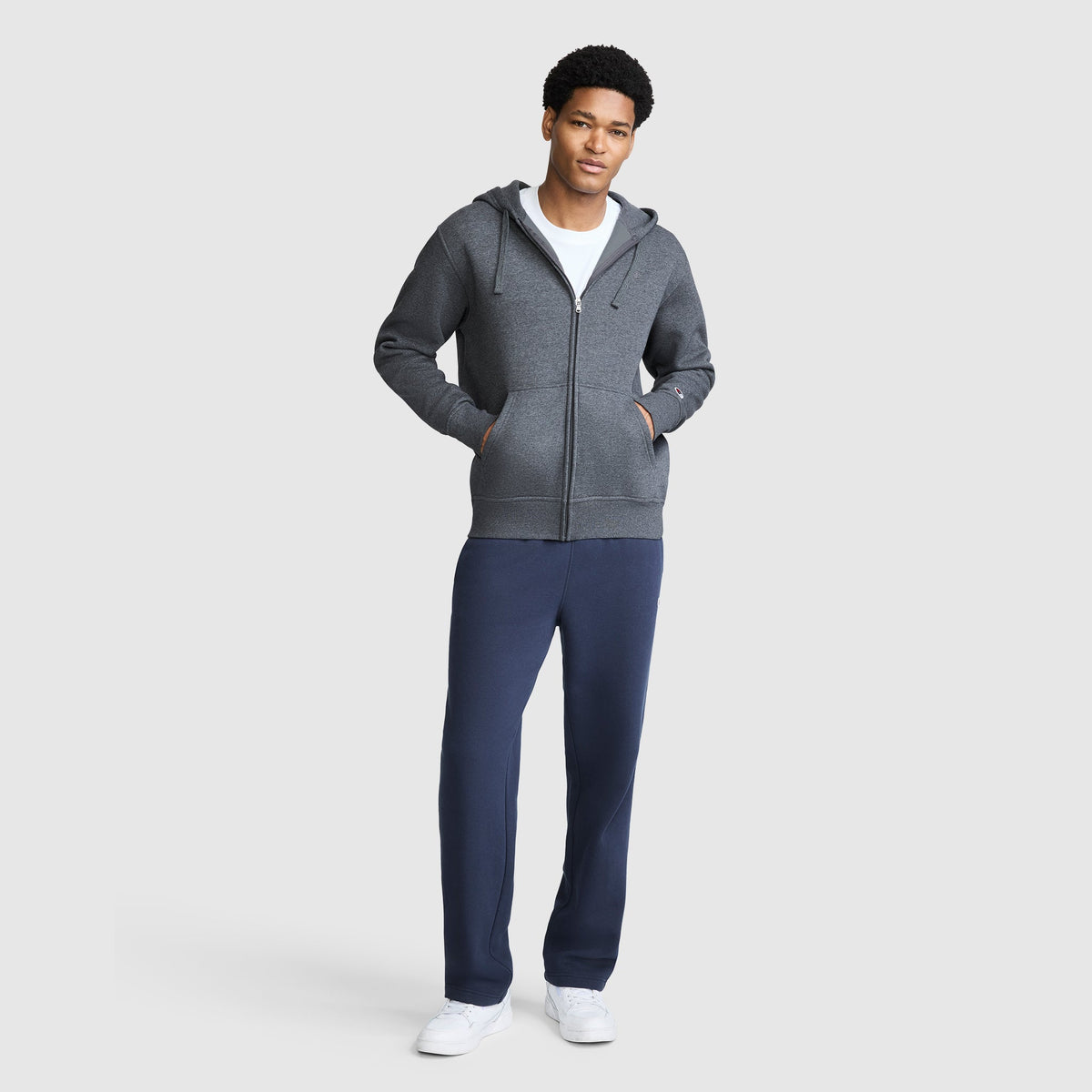 A man stands facing forward, wearing a Champion Powerblend Full-Zip Hoodie with a C Logo in gray over a white shirt, navy blue Champion men's sweatpants with a Powerblend cinched bottom and brushed fleece, and white sneakers. The background is plain and light gray.