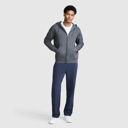 A man stands facing forward, wearing a Champion Powerblend Full-Zip Hoodie with a C Logo in gray over a white shirt, navy blue Champion men's sweatpants with a Powerblend cinched bottom and brushed fleece, and white sneakers. The background is plain and light gray.