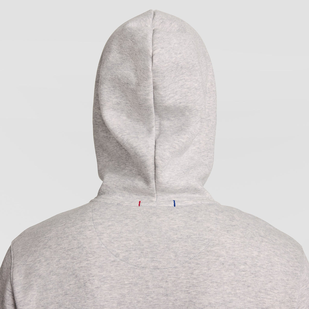 A person wearing a light gray Champion Powerblend Full-Zip Hoodie, C Logo, with the hood up is shown from behind against a plain light background. Two small stitches, one red and one blue, are visible on the back of the men’s sweatshirt.