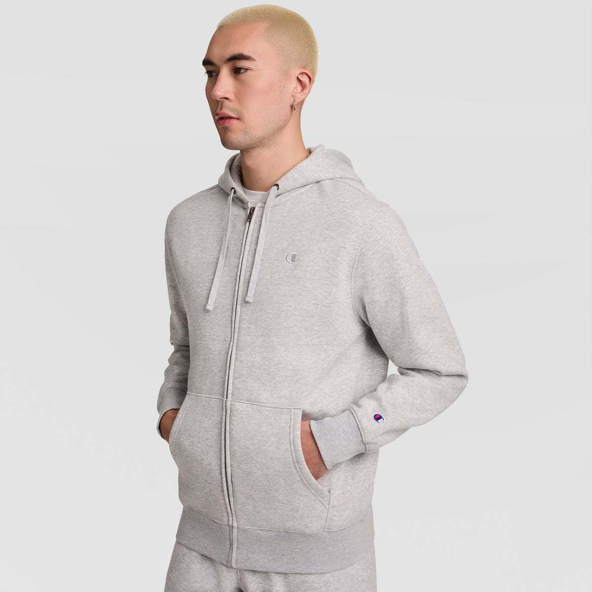 A person with short blond hair wears a light gray Champion Powerblend Full-Zip Hoodie, featuring the C Logo, and matching pants, standing against a plain white background with hands in pockets.