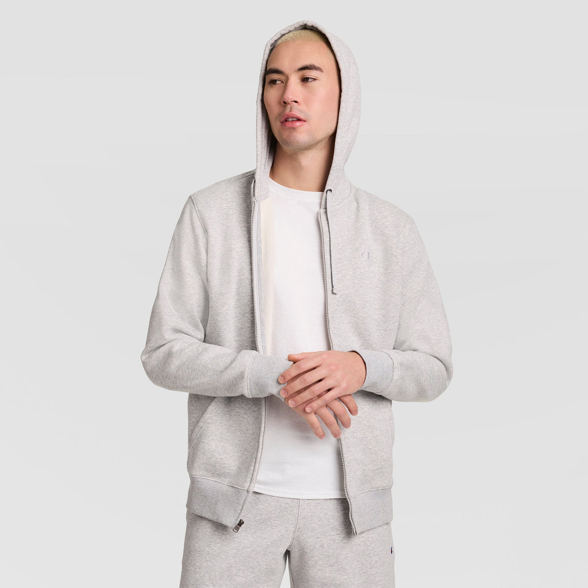A person wearing a light gray Champion Powerblend Full-Zip Hoodie, C Logo, stands against a plain light background, gazing slightly to the side with hands loosely clasped.