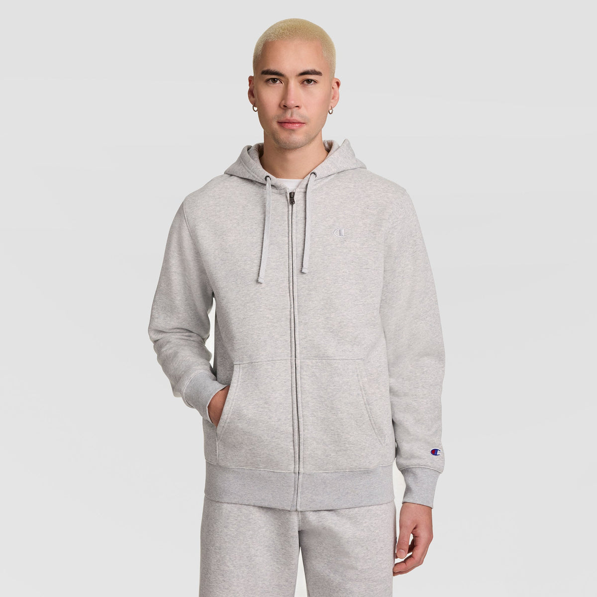 A person with short blond hair stands against a plain white background, wearing a light gray Champion Powerblend Full-Zip Hoodie, C Logo, paired with matching sweatpants and resting one hand in a pocket.