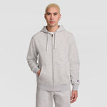 A person with short blond hair stands against a plain white background, wearing a light gray Champion Powerblend Full-Zip Hoodie, C Logo, paired with matching sweatpants and resting one hand in a pocket.