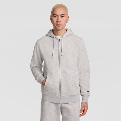 A person with short blond hair stands against a plain white background, wearing a light gray Champion Powerblend Full-Zip Hoodie, C Logo, paired with matching sweatpants and resting one hand in a pocket.