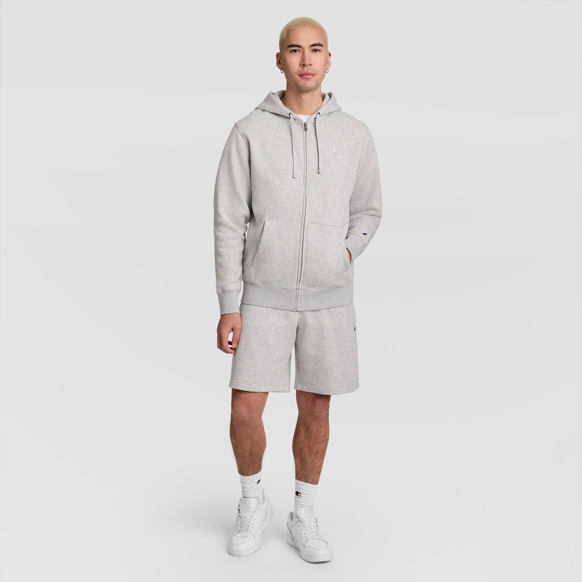 A person stands facing forward, wearing a light gray Champion Powerblend Full-Zip Hoodie, C Logo, paired with matching shorts, white socks, and white sneakers against a plain white background.