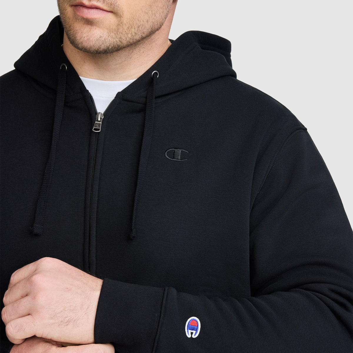 A man wearing a black Champion Powerblend Full-Zip Hoodie, C Logo – Big & Tall, featuring drawstrings and signature small Champion logos on the chest and wrist, stands against a plain light background.