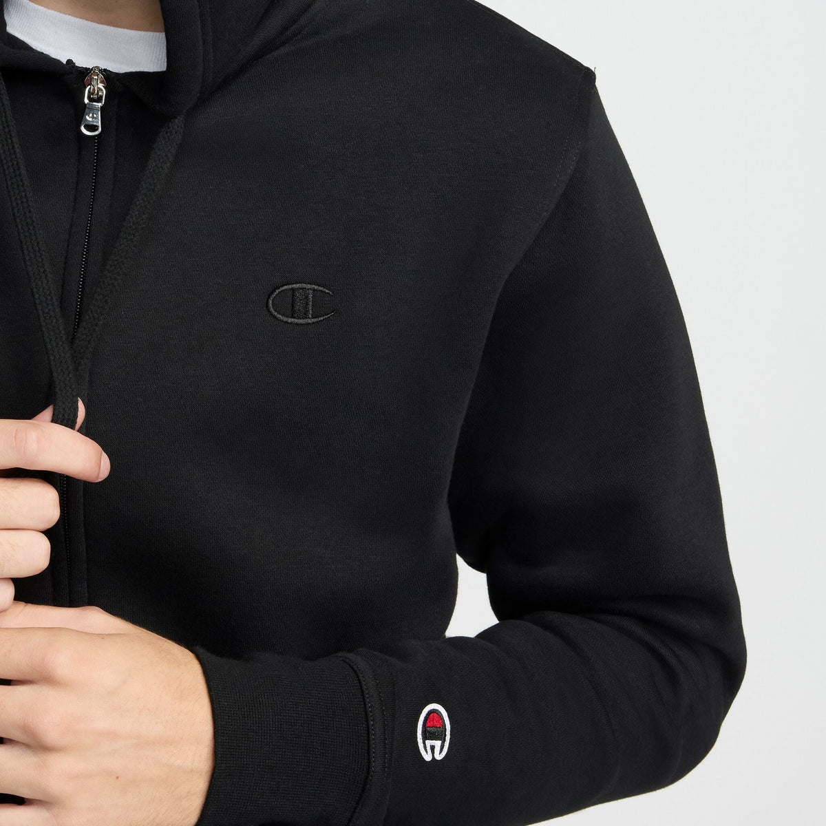 A person wearing the Champion Powerblend Full-Zip Hoodie, C Logo in black, featuring a small Champion logo on the chest and sleeve, partially unzipping the jacket.