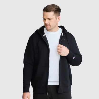 A man wearing a black Champion Powerblend Full-Zip Hoodie, C Logo - Big & Tall, layered over a white t-shirt, stands against a light gray background, looking to his right with one hand touching his chest.
