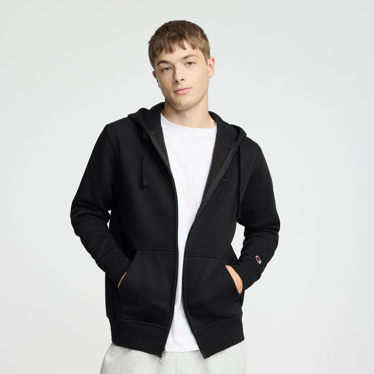 A young man wearing a Champion Powerblend Full-Zip Hoodie, C Logo, over a white t-shirt and light gray pants stands with his hands in his pockets against a plain light background.