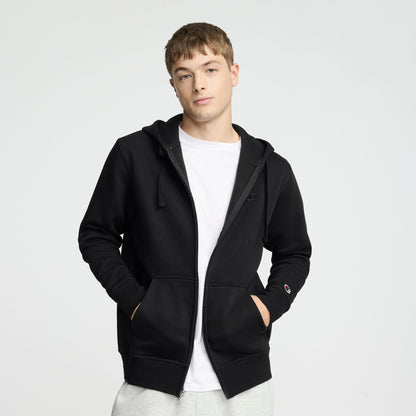A young man wearing a Champion Powerblend Full-Zip Hoodie, C Logo, over a white t-shirt and light gray pants stands with his hands in his pockets against a plain light background.