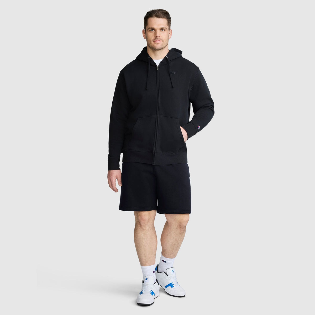 A man stands facing forward, wearing a Champion Powerblend Full-Zip Hoodie, C Logo - Big & Tall in black, paired with black shorts, white socks, and white sneakers with blue accents, all set against a plain light gray background.