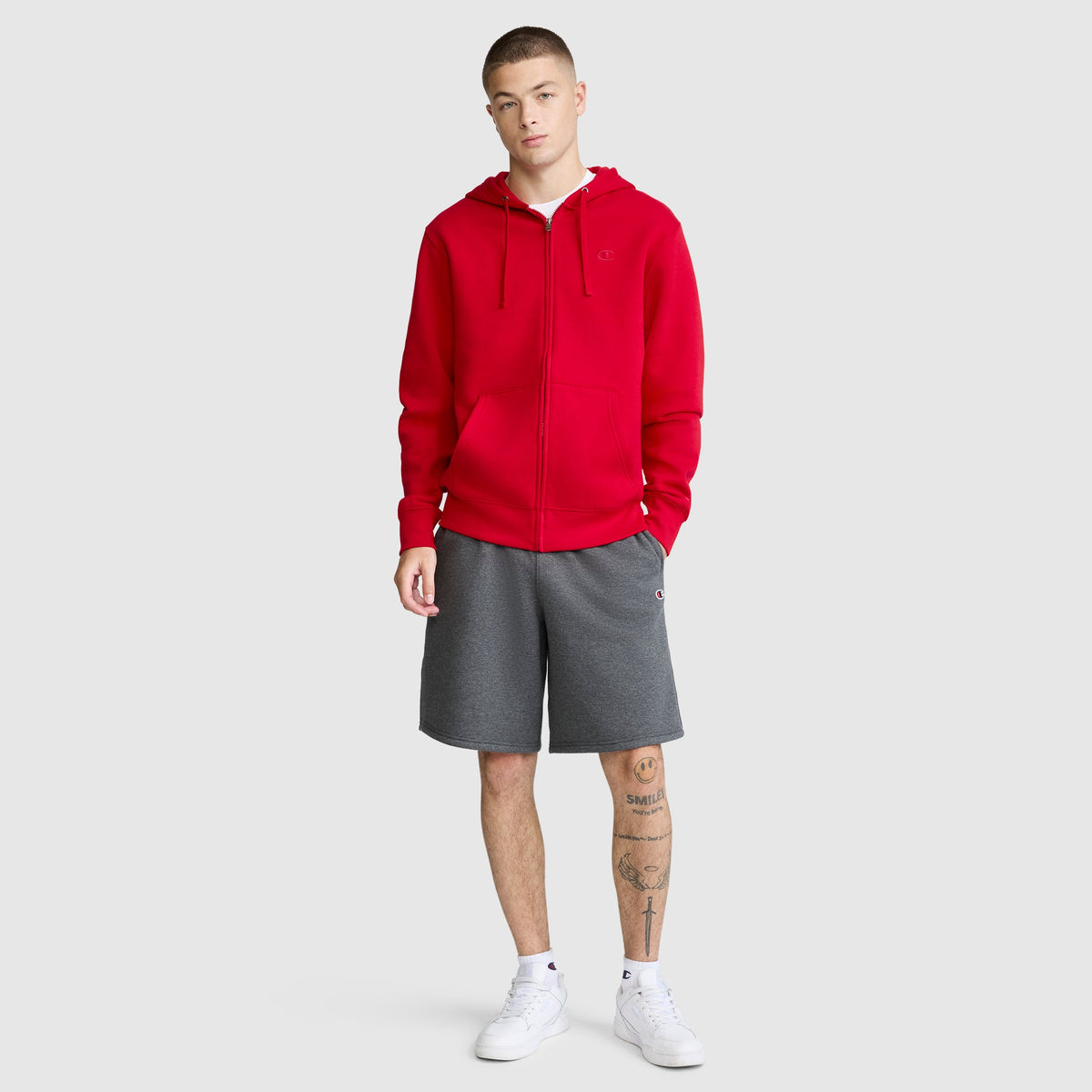 A person wearing the Champion Powerblend Full-Zip Hoodie with a C logo in red, paired with gray men's fleece sweatpants featuring a cinched bottom and white sneakers, stands facing forward. The individual has short hair and visible leg tattoos. The background is plain and light gray.