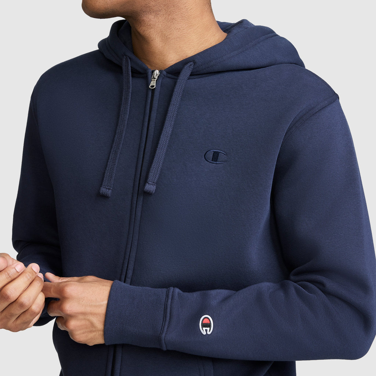 A person wearing a navy blue Champion Powerblend Full-Zip Hoodie, C Logo, with a small embroidered logo on the chest and a patch on the left sleeve.
